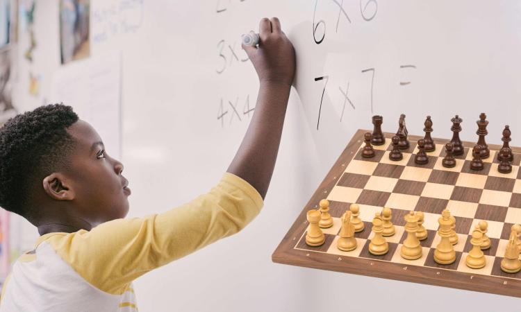 Barn räknar matte på tavla bredvid ett schackbräde. 